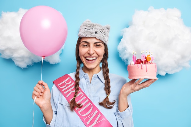 mulher com duas tranças gosta de festa de aniversário segura bolo de morango balão de hélio inflado se diverte expressa emoções positivas comemora aniversário em casa