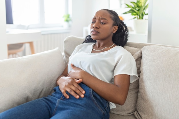 Mulher com expressão dolorosa de mãos dadas contra a barriga, sofrendo de dor menstrual, deitada triste na cama de casa, tendo cãibras de barriga no conceito de saúde feminina