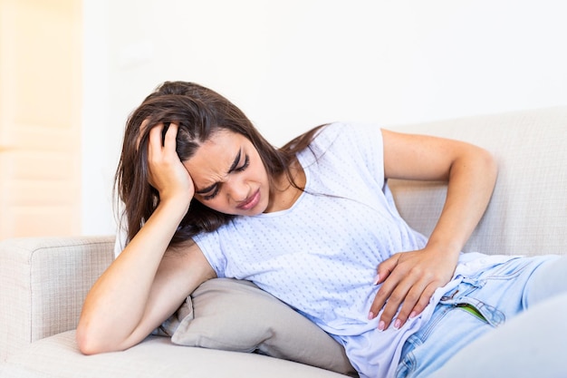 Foto mulher com expressão dolorosa segurando as mãos contra a barriga sofrendo dor do período menstrual deitada triste na cama de casa tendo cãibra na barriga no conceito de saúde feminina