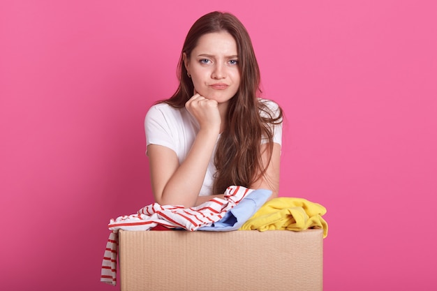 mulher com expressão facial pensativa, posando perto da caixa com roupas velhas, decide quem dar as coisas para uso secundário, em cima de rosa. Conceito de doação.