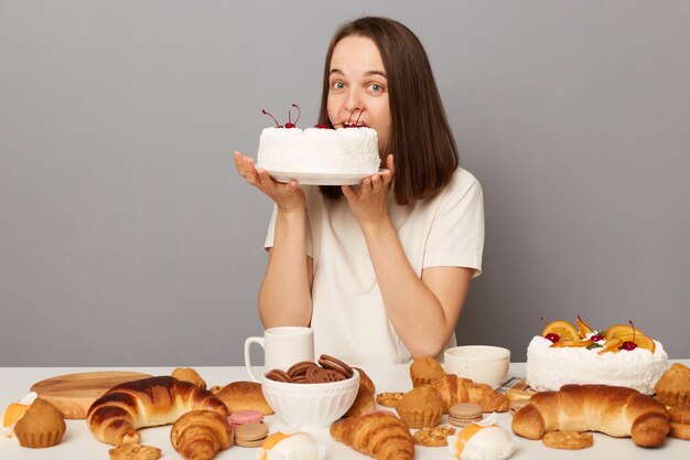 Mulher com fome vestindo camiseta branca isolada sobre fundo cinza mordendo bolo grande sentado à mesa com confeitaria olhando para a câmera com expressão facial engraçada