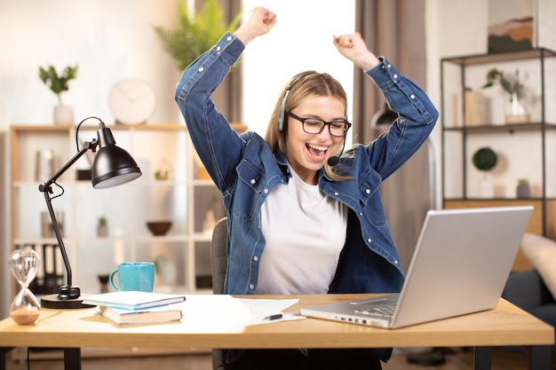 Mulher com fone de ouvido e óculos sentada com os braços levantados e sorrindo enquanto olha para a tela do laptop