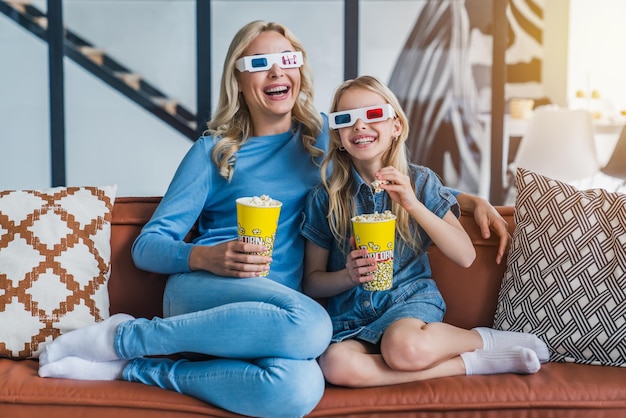 Mulher com garotinha em óculos 3d assistindo filme e comendo pipoca em casa