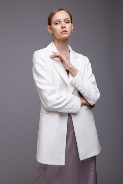 Mulher com jaqueta branca longa saia metálica prateada posando em fundo cinza Retrato de estúdio