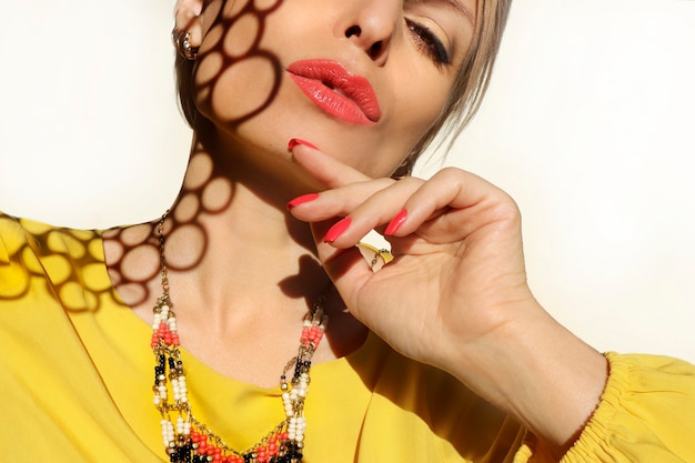 Mulher com lábios cor de coral e unhas em um vestido amarelo sobre um fundo claro com um padrão de sombra no rosto.