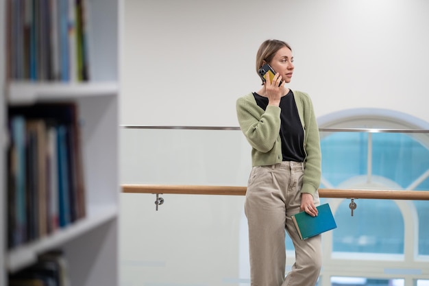 Mulher com livro nas mãos falando no celular na biblioteca pública moderna