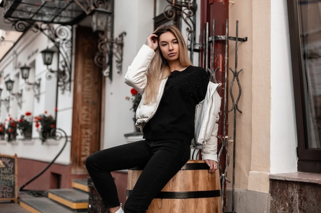 Foto mulher com longos cabelos loiros e uma jaqueta branca da moda sentada em um barril vintage de madeira