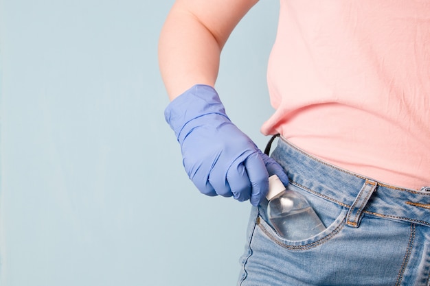 Mulher com luvas de borracha azul colocando gel desinfetante no bolso da frente da calça jeans