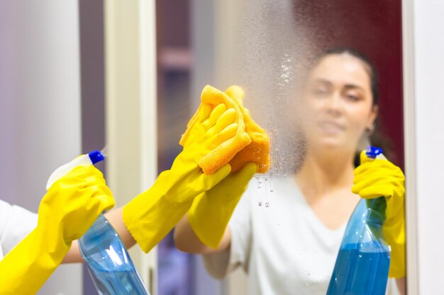 Mulher com luvas de borracha pulveriza detergente e limpa o espelho com trapo Vista do ombro Serviço de limpeza e tarefas