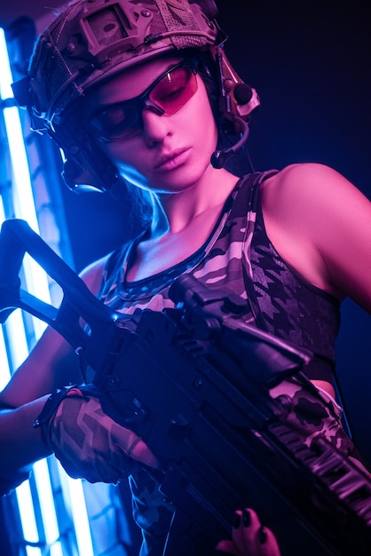 Mulher com macacão militar de airsoft posando com uma arma nas mãos em uma parede escura no meio da névoa sob a luz de neon