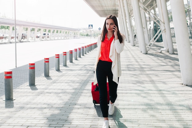 Mulher com mala vermelha anda ao longo do aeroporto e fala no telefone