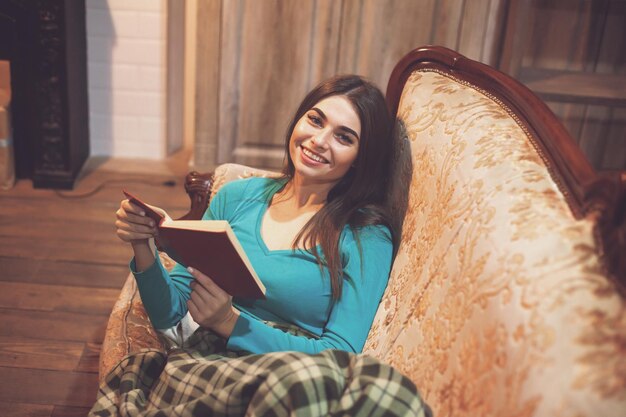 Mulher com manta está lendo um livro vermelho no sofá confortável