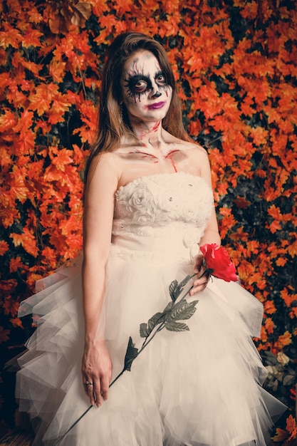 Mulher com maquiagem de fantasma e vestido de noiva segurando uma rosa.