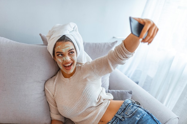 Mulher com máscara de argila tomando selfie com telefone celular em casa desfrutando de relaxamento