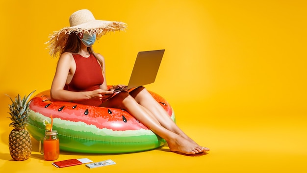 Mulher com máscara e chapéu sentada em um anel inflável enquanto segura um cartão de crédito e faz compras on-line com laptop
