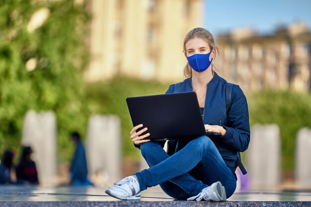Mulher com máscara facial protetora e laptop sentada do lado de fora