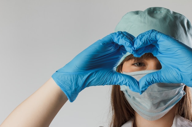 Mulher com máscara médica e mãos em luvas de látex mostra o símbolo do coração. Corona de proteção