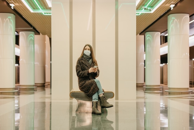 Mulher com máscara médica para evitar a disseminação do COVID está sentada com um smartphone e esperando o trem do metrô. Uma garota de cabelo comprido com máscara cirúrgica mantém distância social no metrô.