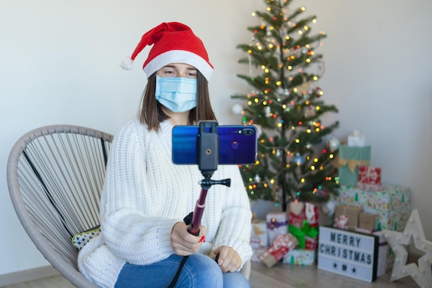 Mulher com máscara médica perto da árvore de natal fazendo uma videochamada no telefone