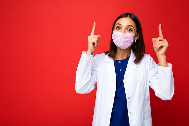 Mulher com máscara protetora de vírus no rosto contra coronavírus e jaleco branco médico isolado no fundo e apontando os dedos para cima.