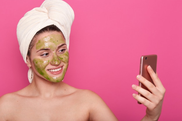 Mulher com máscara verde no rosto, toalha branca enrolada, posa seminua, tomando selfie no smartphone no banheiro