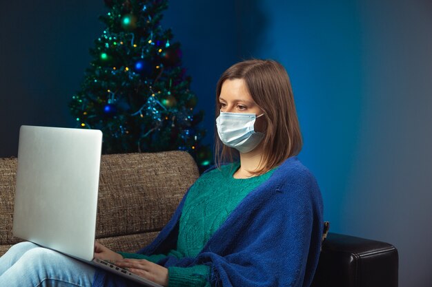 Mulher com notebook com máscara protetora, sentada no sofá com árvore de natal decorada