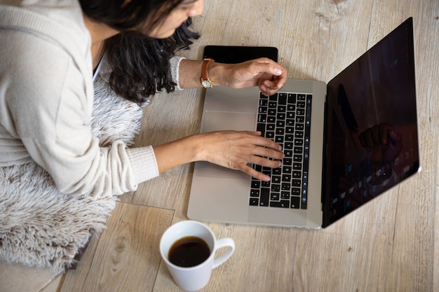 Mulher com o laptop on-line em casa tomando café, com telefone celular