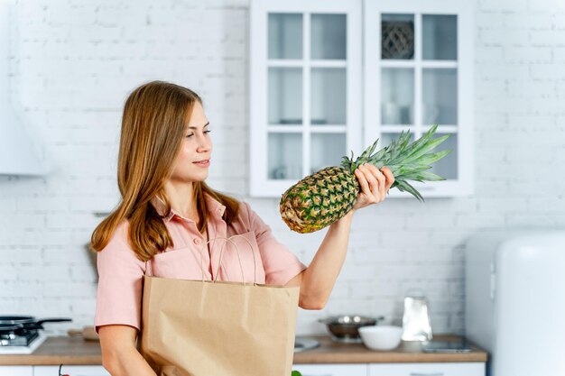 Mulher com o pacote de mercearia nas mãos Fundo da cozinha Jovem mulher com comida saudável Abacaxi nas mãos da jovem