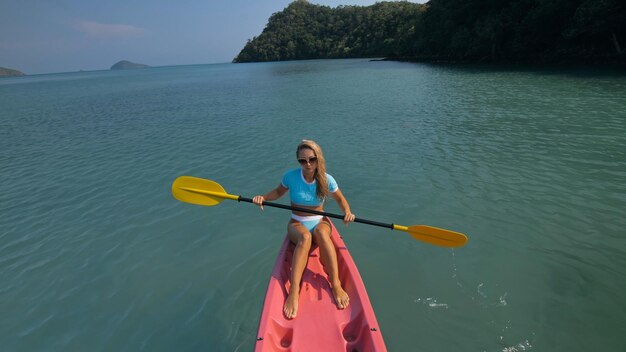 Mulher com óculos de sol enfileira canoa de plástico rosa ao longo da água do mar contra colinas verdes e céu azul