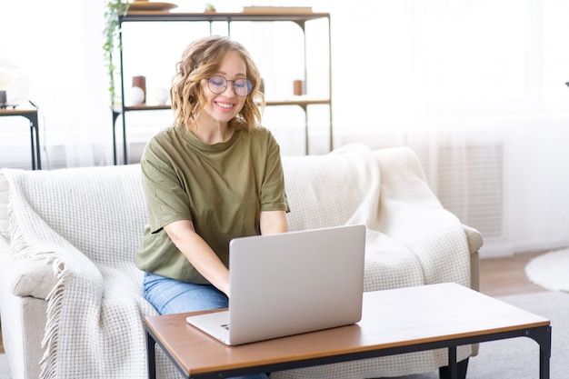 Mulher com óculos usa laptop digitando teclado sentado no sofá janela grande fundo interior da casa mulher autônoma trabalhando em casa
