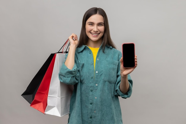 Mulher com olhar satisfeito mostrando sacolas de compras e smartphone com tela em branco para propaganda