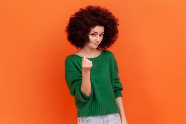 Mulher com penteado afro no suéter verde esfregando os dedos mostrando gesto de dinheiro, pedindo salário, exigindo suborno, olhando para a câmera com um sorriso. Tiro de estúdio interior isolado em fundo laranja.