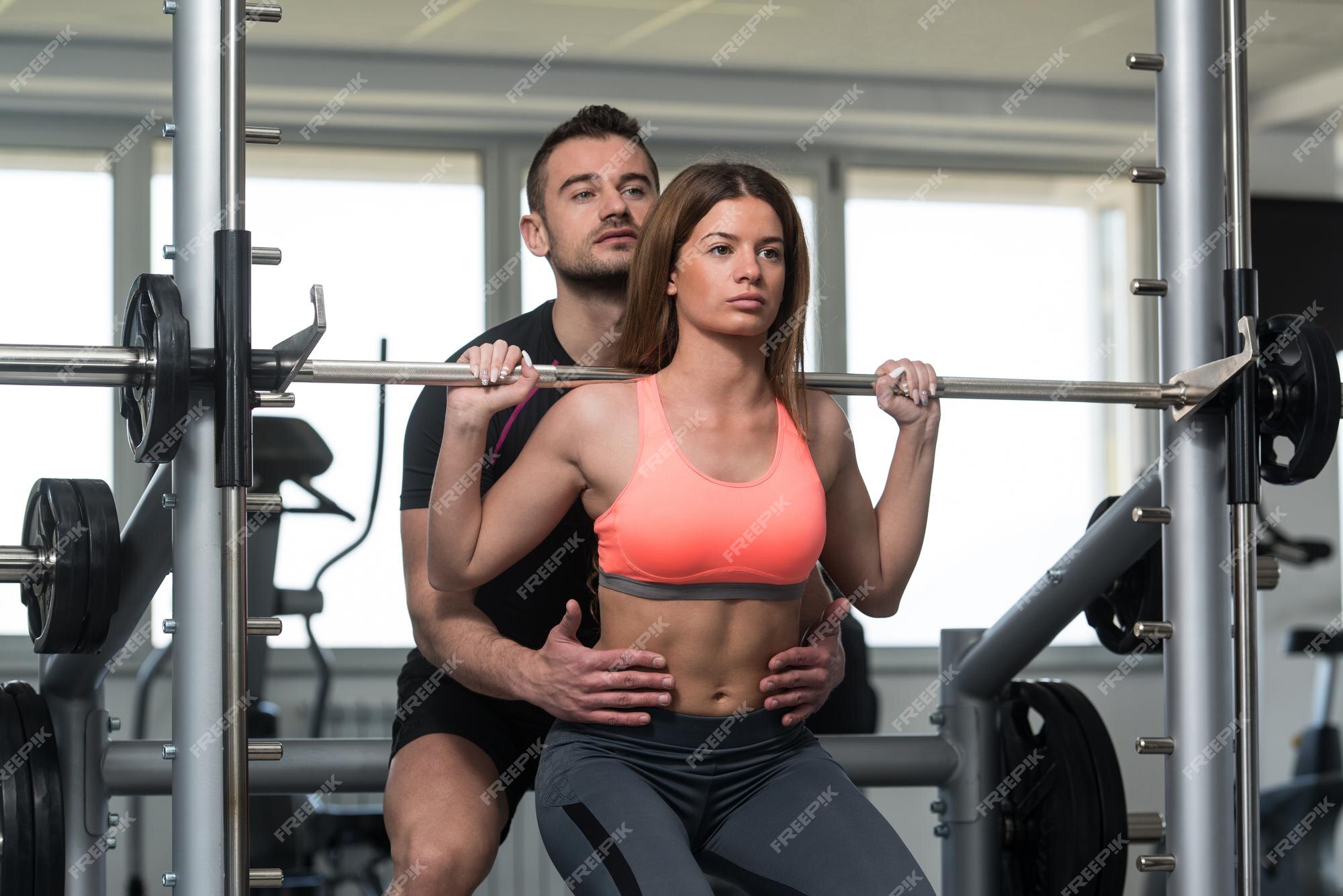 Uma foto de corpo inteiro da mulher esportiva que está subindo a barra para  fazer agachamentos na prateleira de agachamento em uma academia. uma menina  morena musculosa usando um top coral e