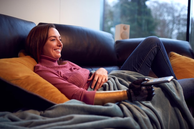 Mulher com prótese de braço e mão em casa relaxando no sofá assistindo TV