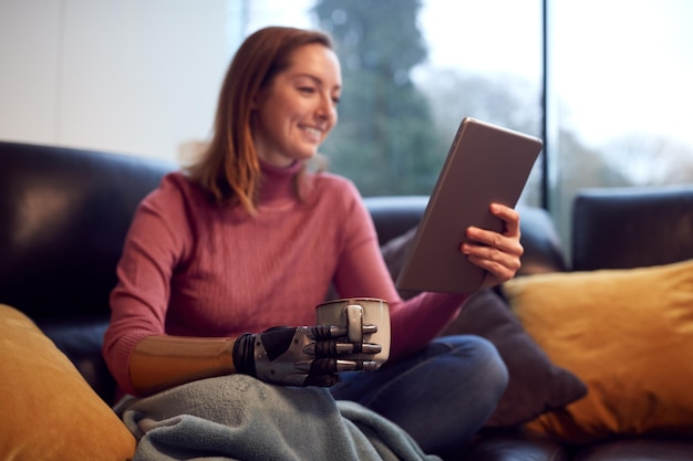 Mulher com prótese de braço e mão em casa usando tablet digital bebendo café