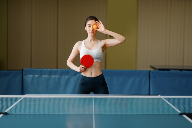 Mulher com raquete mostra bola de pingue-pongue dentro de casa.