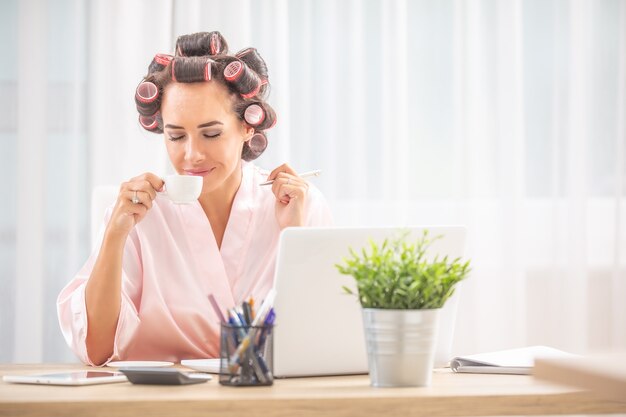 Mulher com rolos de cabelo e camisola cheira café enquanto está sentada na frente do computador em casa.