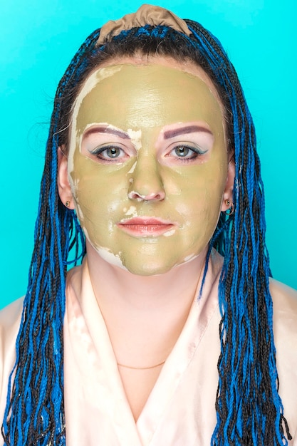 Mulher com rosto de tranças afro azuis em uma máscara feita de argila verde sobre fundo azul. Foto vertical