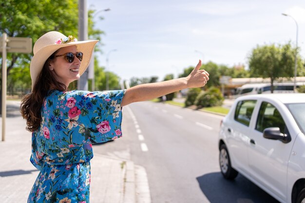 Mulher com roupas de verão e chapéu pedindo carona em uma estrada urbana