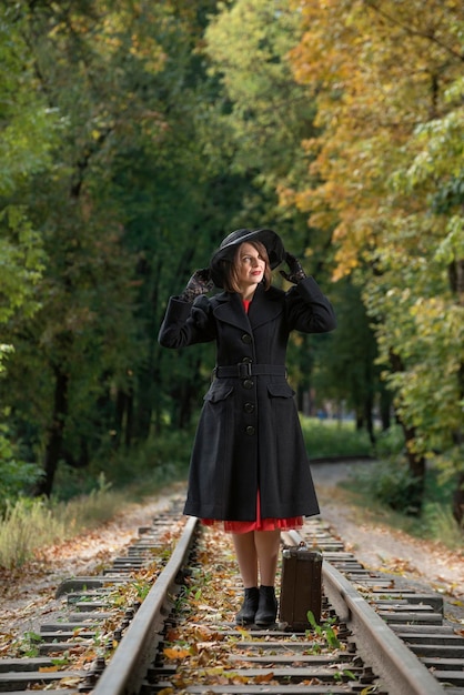 Mulher com roupas retrô casaco preto e chapéu de aba larga fica com valise velha nos trilhos Mulher vestida no estilo dos anos 40