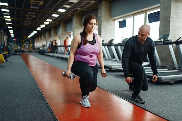 Mulher com sobrepeso segura halteres, exercício com treinador no clube desportivo, treino de ajuste com instrutor