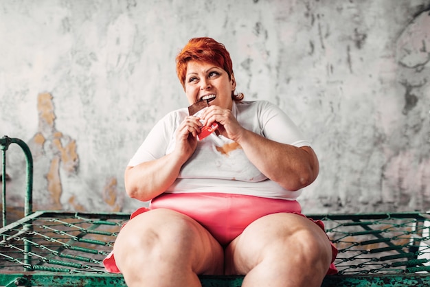 Mulher com sobrepeso senta na cama e come chocolate, preguiça e obesidade, bulímica. Comer alimentos não saudáveis, mulher gorda