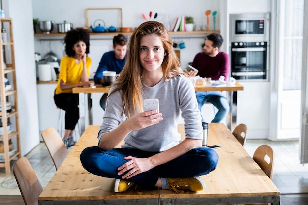 Mulher com telefone celular sentada na mesa de jantar em casa com amigos ao fundo