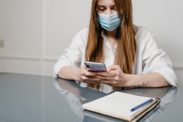 Mulher com telefone e notebook em casa na mesa com máscara