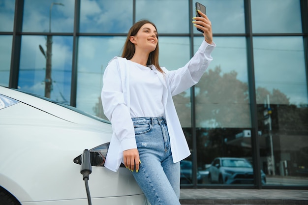 Mulher com telefone perto de um carro elétrico alugado veículo carregado na estação de carregamento