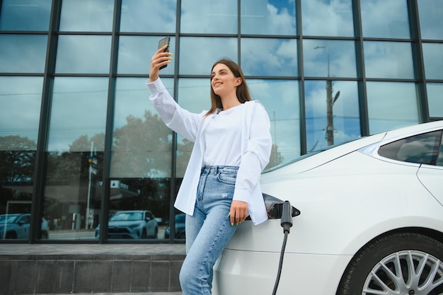 Mulher com telefone perto de um carro elétrico alugado Veículo carregado na estação de carregamento