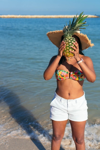 Mulher com um abacaxi no rosto caminhando na praia