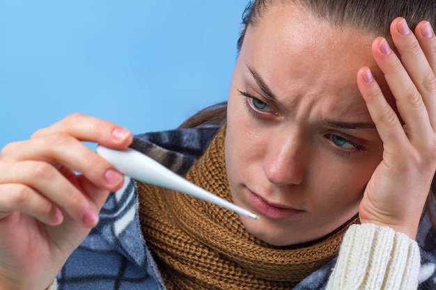 Mulher com um cachecol de malha e um pulôver segura um termômetro nas mãos e se sente mal