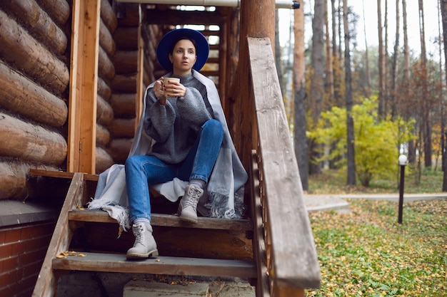 Mulher com um chapéu azul e um lenço e uma caneca está em uma casa de madeira na floresta