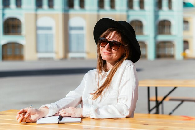 Mulher com um chapéu e óculos escuros sorrindo enquanto está sentada em um café pensa nas anotações para escrever em um caderno